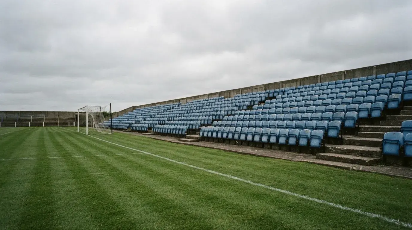 Rzędy pustych plastikowych krzesełek małego stadionu widziane z poziomu murawy pod pochmurnym niebem.