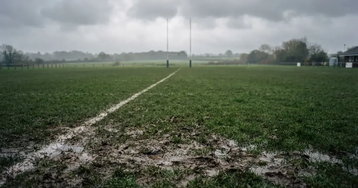 Boisko do rugby ze słupami bramki w oddali pod pochmurnym niebem, mokra trawa i ślady butów.